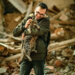 A man wearing glasses joyfully holds a tabby cat in a dilapidated urban setting.